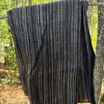 A dark charcoal gray knit blanket with vertical beige and white stripes, hanging from a tree branch outdoors in a wooded setting.