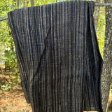 A dark charcoal gray knit blanket with vertical beige and white stripes, hanging from a tree branch outdoors in a wooded setting.