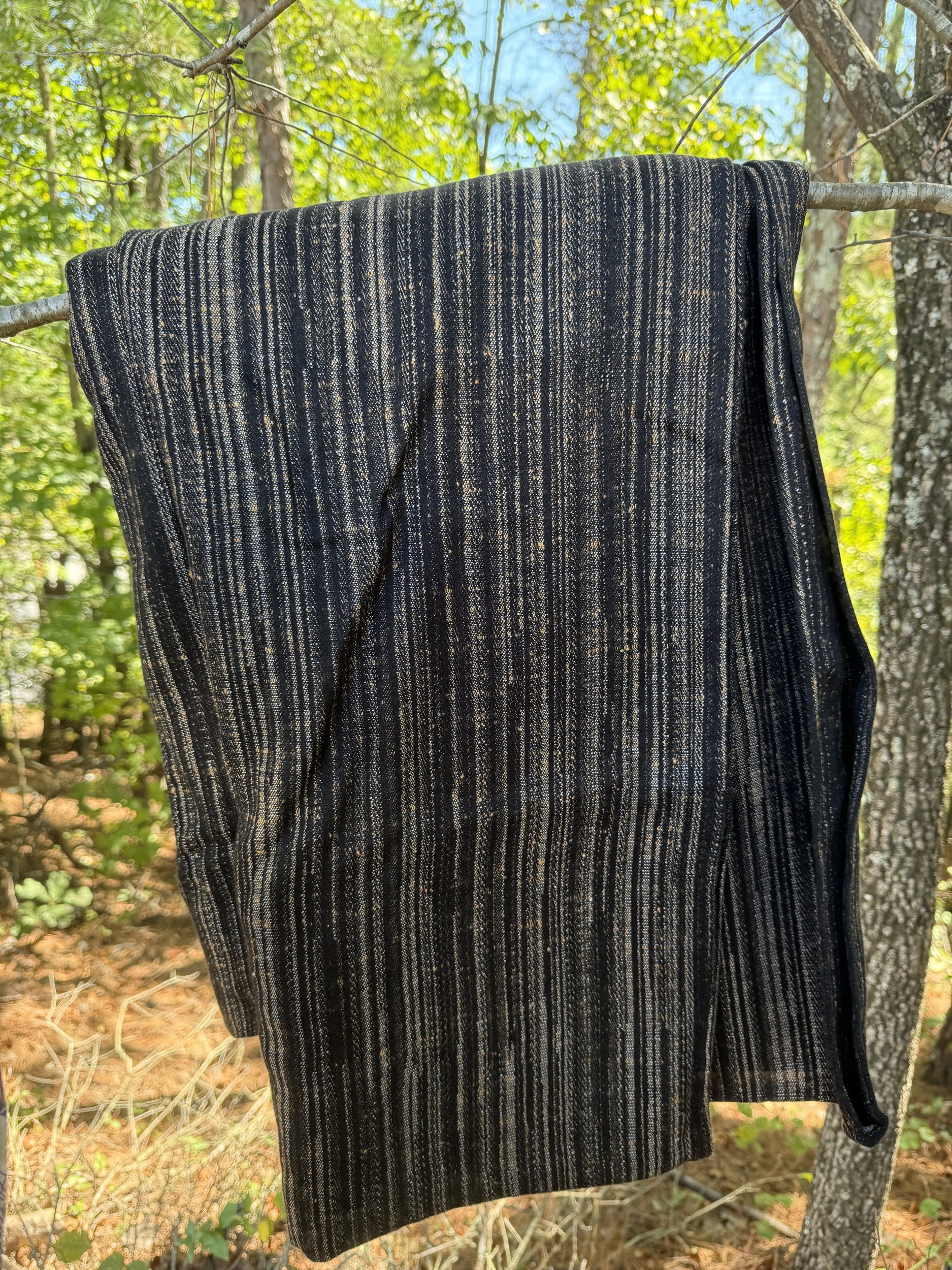 A dark charcoal gray knit blanket with vertical beige and white stripes, hanging from a tree branch outdoors in a wooded setting.