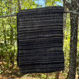 A dark woven blanket with horizontal stripes of black, navy, and subtle tan flecks, hanging from a tree branch in a forest setting.
