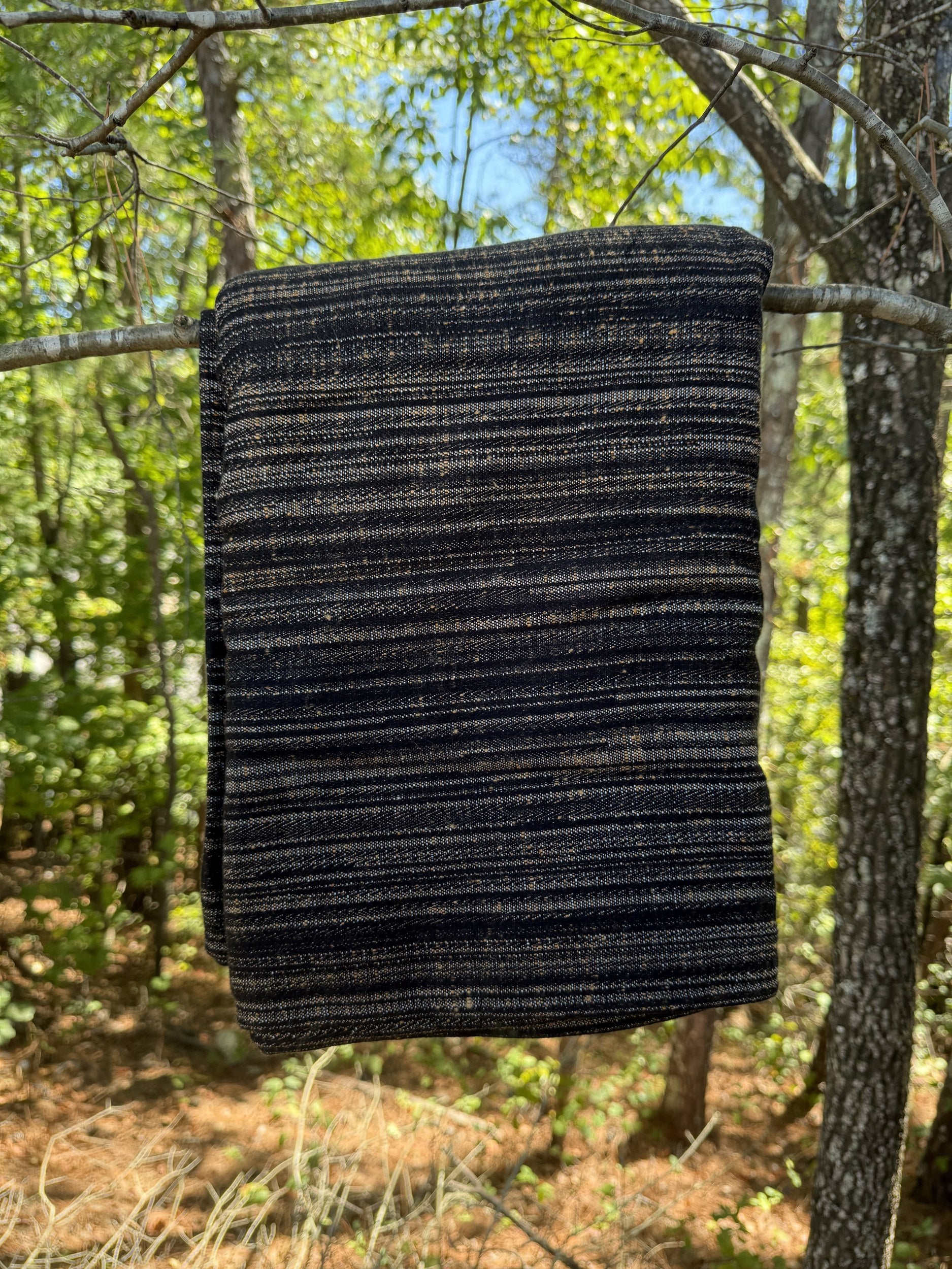 A dark woven blanket with horizontal stripes of black, navy, and subtle tan flecks, hanging from a tree branch in a forest setting.
