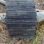 A folded woven textile, likely a blanket, throw, or scarf, featuring vertical stripes in black and natural tan, displayed outdoors on a tree trunk surrounded by pine needles and forest floor debris.