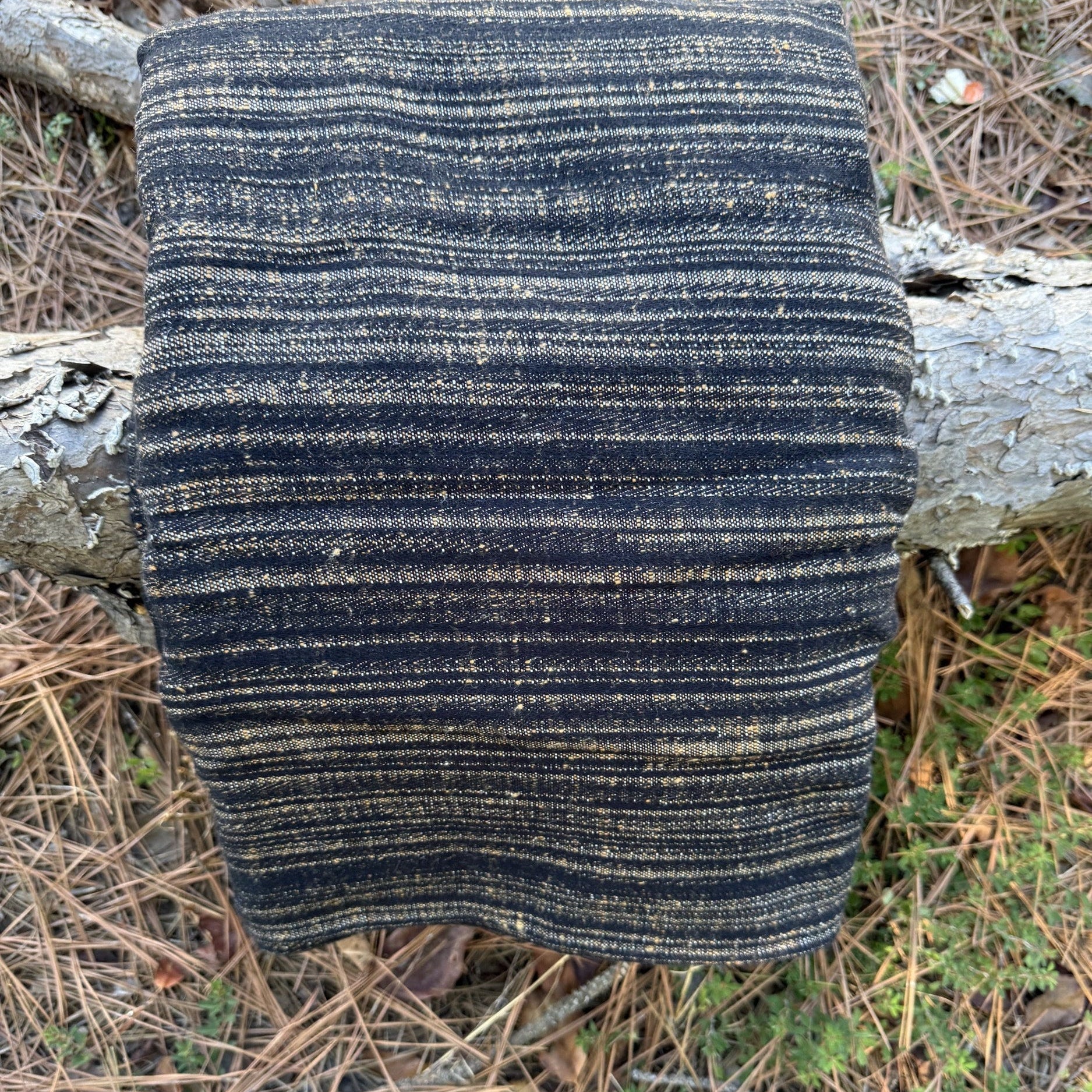 A folded woven textile, likely a blanket, throw, or scarf, featuring vertical stripes in black and natural tan, displayed outdoors on a tree trunk surrounded by pine needles and forest floor debris.