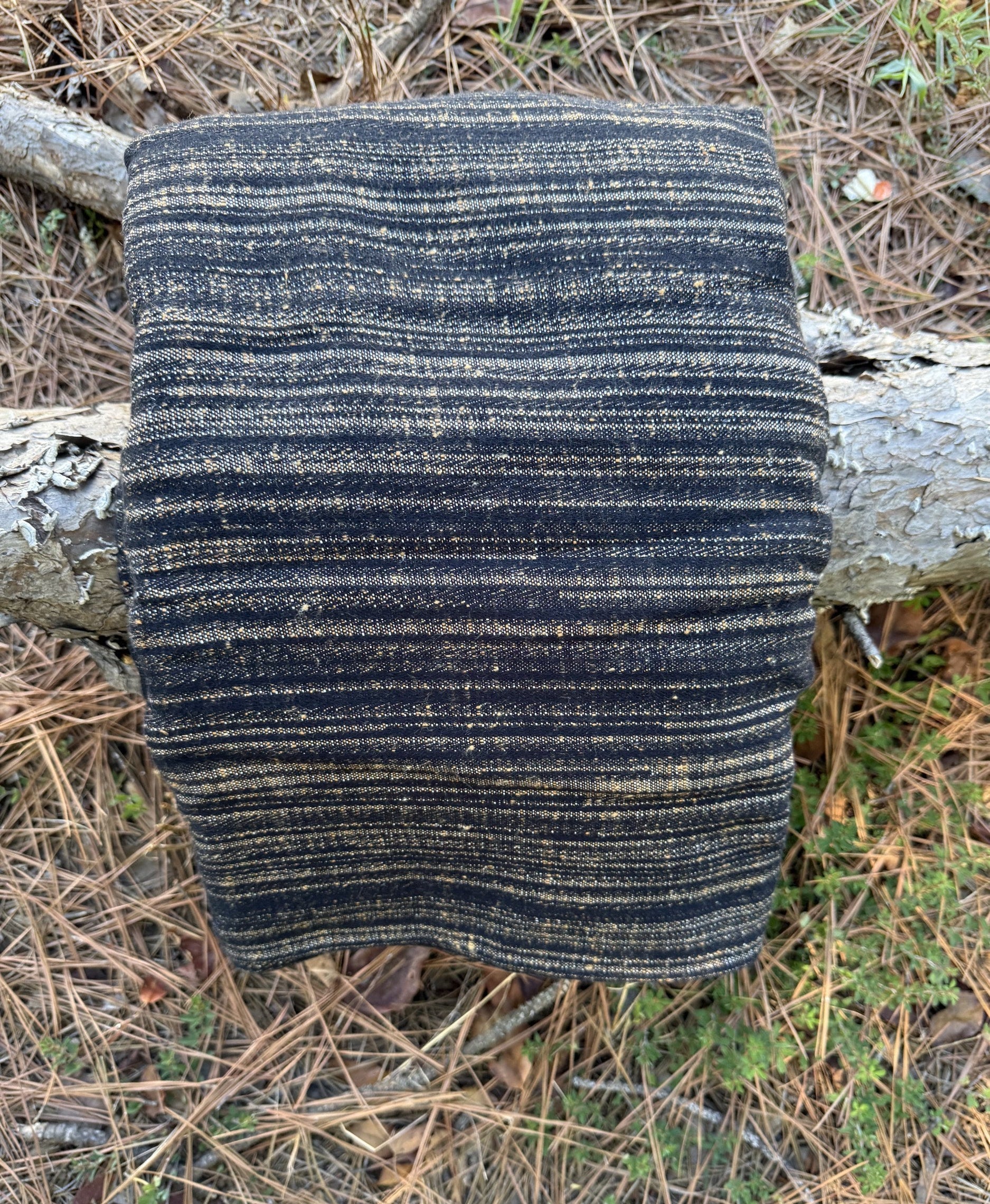 A folded woven textile, likely a blanket, throw, or scarf, featuring vertical stripes in black and natural tan, displayed outdoors on a tree trunk surrounded by pine needles and forest floor debris.