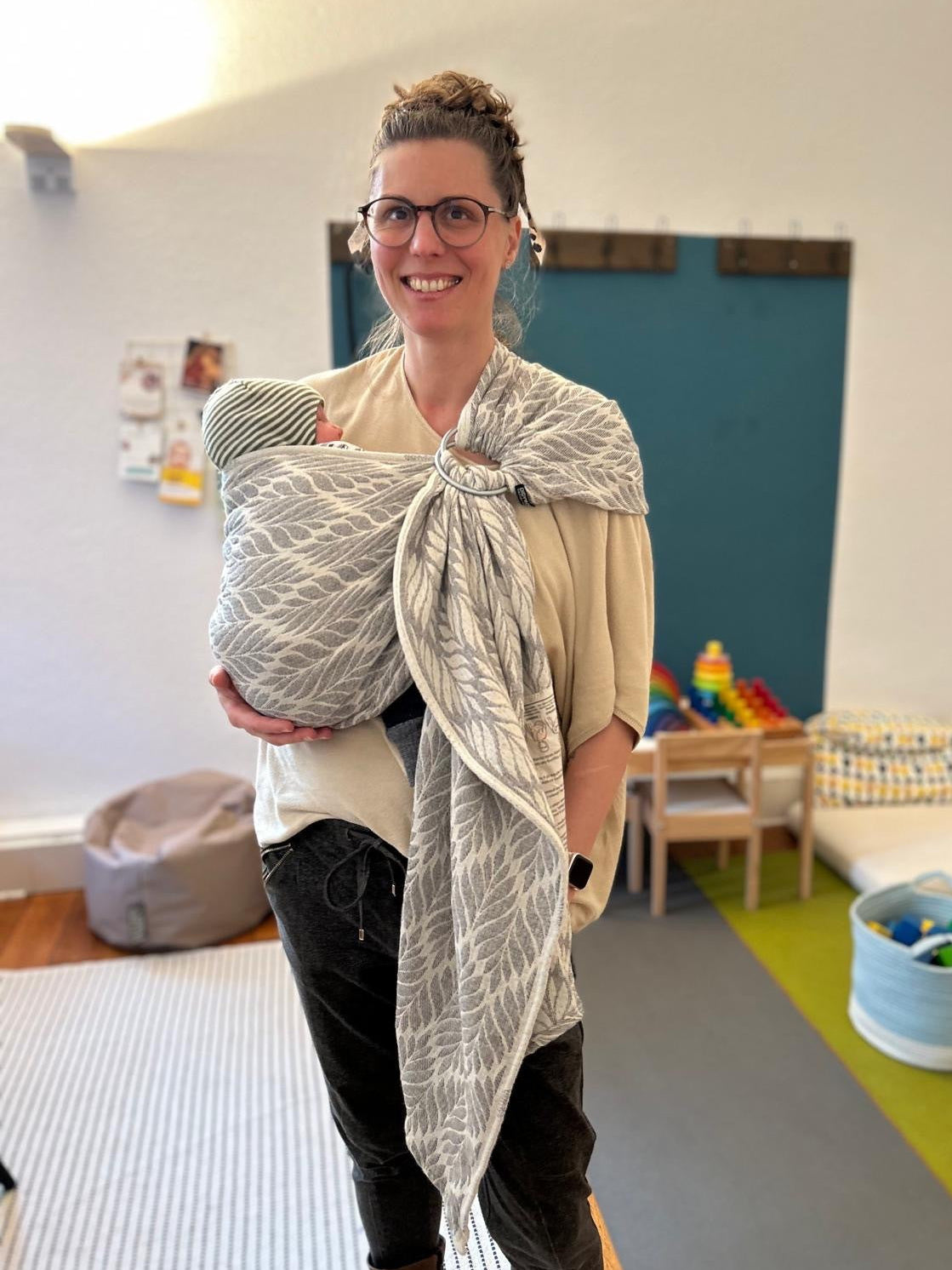Woman holding a baby in a sling in a room with toys and furniture.
