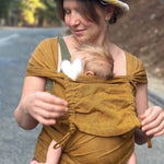 A woman wearing a straw hat and a mustard yellow baby wrap carrier with a baby inside, standing outdoors on a road with trees in the background.
