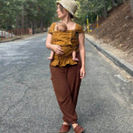 A woman wearing a mustard yellow brown baby wrap carrier  and brown pants stands on an asphalt road surrounded by trees, holding a baby in a woven straw hat and baby carrier outfit.