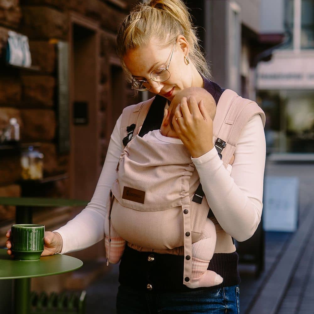 Woman holding a baby in a sling and a green mug outdoors.