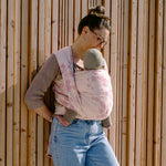 Woman holding a baby in a floral baby carrier against a wooden wall.