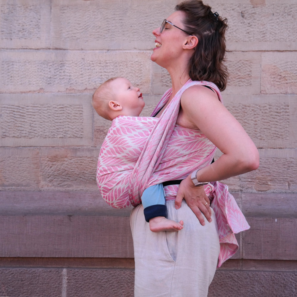 A woman wearing glasses carries a baby in a pink and white patterned wrap carrier against a stone wall background.